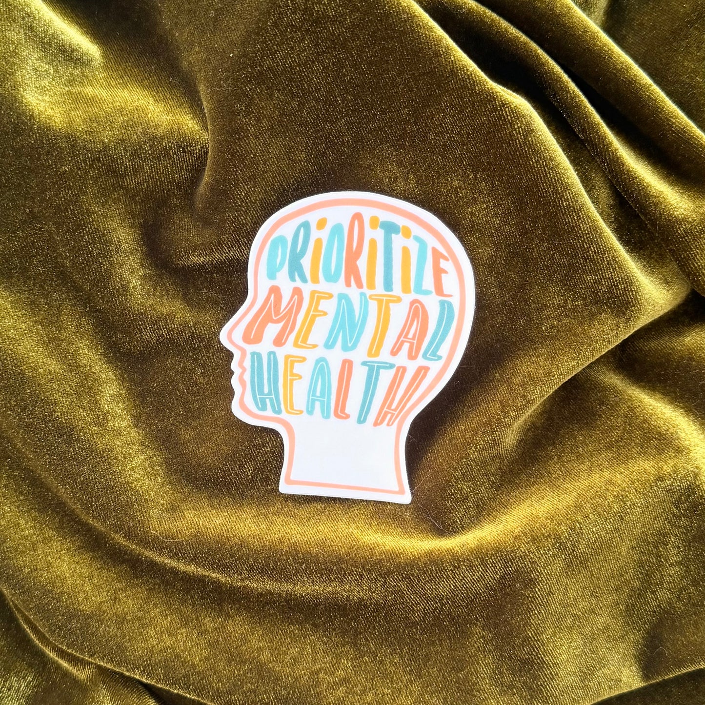 Sticker with 'Prioritize Mental Health' text on a textured green fabric background